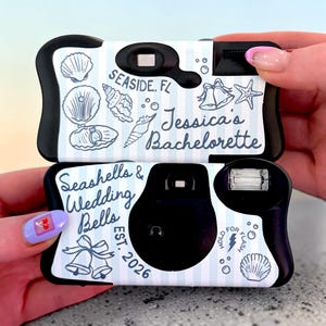 May include: Two black disposable cameras with white and blue seashell and wedding bell designs. The top camera reads "Seaside, FL" and "Jessica's Bachelorette." The bottom camera reads "Seashells & Wedding Bells EST. 2026."
