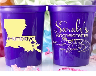 May include: Two purple plastic cups. One cup features a yellow outline of Louisiana with the text "#Humblaya". The other cup has the text "Sarah's Bachelorette New Orleans 2020" with an alligator illustration.