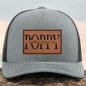 May include: A gray trucker hat with a black mesh back and a curved brim. The hat features a brown leather patch with the word "POPPY" and the names "Charlotte & Harper & Ezra" in black lettering.