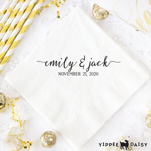 May include: White paper napkin with black calligraphy text that reads "emily & jack" and "NOVEMBER 21, 2020". The napkin is on a white lace tablecloth with gold confetti and gold-colored ornaments.