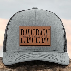 May include: Gray trucker hat with a black mesh back and a brown leather patch. The patch features the text "DAIA/DAIA" above "Alexander & Houston & Axel" and "IAIA/IAIA" below.