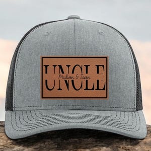 May include: A gray trucker hat with a black mesh back. The front features a brown leather patch with the word "UNCLE" in bold, black letters, and the names "Madison & Jason" below. A stylish accessory.