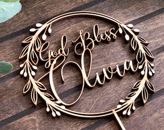 May include: Wooden cake topper with a floral wreath design. The topper features the text "God Bless Olivia" in a cursive font.