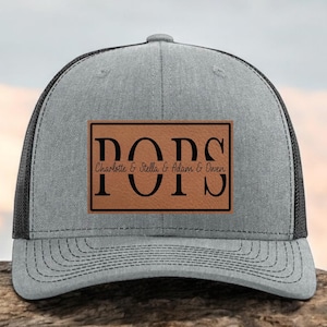 May include: A gray trucker hat with a black mesh back and a curved brim. The hat features a brown rectangular patch with the word "POPS" and the names "Charlotte & Stella & Adam & Owen" in black lettering.