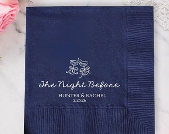 May include: Navy blue cocktail napkin with white text and a decorative design of two champagne glasses. The text reads "The Night Before" with a couple's names and a date. Pink roses are in the background.