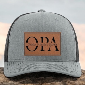 May include: A gray trucker hat with a black mesh back and a brown leather patch. The patch features the word "OPA" in large black letters, with the names "Christian & Beatrix & Charlotte" below.