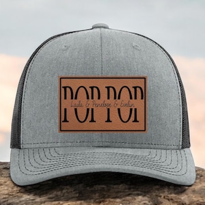 May include: Gray trucker hat with a black mesh back and a brown rectangular patch. The patch features the word "DAD" in a mirrored design above the names "Laila & Penelope & Evelin" and the word "IUTU" in a mirrored design.