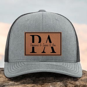 May include: A gray trucker hat with a black mesh back. The front features a brown rectangular patch with the letters "DA" and the names "Dominic & Isaac & Ava" in black text. The hat is on a wooden surface.