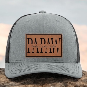 May include: Gray trucker hat with a black mesh back and a curved brim. The hat features a brown leather patch with the text "DA DA DA" above the names "Amelia & Luke & Caleb" and "DA DA DA" below.