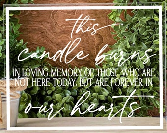May include: A clear acrylic sign with white script that reads "This candle burns in loving memory of those who are not here today, but are forever in our hearts." The sign is surrounded by green foliage.