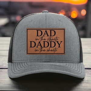 May include: Gray trucker hat with a black mesh back and a brown leather patch. The patch features the text "DAD in the streets DADDY in the sheets" in black lettering. The hat is sitting on a dark wooden surface.
