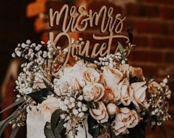 May include: A three-tiered wedding cake decorated with white roses and baby's breath. The cake is topped with a wooden sign that says "Mr. & Mrs. Joucet".