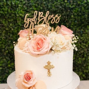 May include: A white two-tiered cake decorated with pink roses, white flowers, and macarons. The cake has a gold cross topper and a wooden sign that says "God Bless Avery".