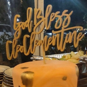 May include: A peach-colored cake with gold accents and a wooden cake topper that says "God Bless Clementine".