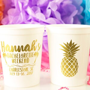 May include: Two white plastic cups with gold lettering. One cup says "Hannah's Bachelorette Weekend Charleston, SC July 13-16, 2017". The other cup has a gold pineapple design.