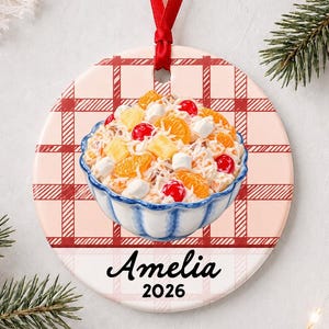 May include: A round ceramic ornament featuring a red plaid background and a central illustration of a bowl filled with fruit salad containing mandarin oranges, pineapple, cherries, and marshmallows. The name Amelia and the year 2026 are printed below.