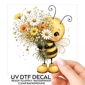 May include: A UV DTF decal featuring a cartoon bee with black and yellow stripes, large eyes, and antennae, holding a large bouquet of white, yellow, and orange wildflowers. The text below reads UV DTF DECAL READY TO APPLY WATERPROOF CLEAR BACKGROUND.