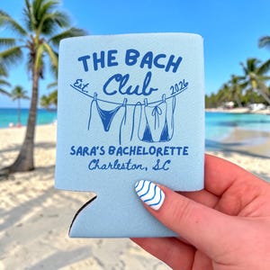 May include: A light blue foam can cooler featuring dark blue text that reads THE BACH CLUB Est. 2026, SARA'S BACHELORETTE Charleston, SC, with a graphic of two bikini tops hanging on a clothesline between the text elements.