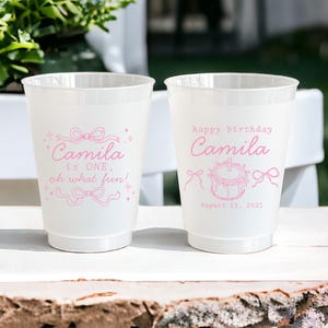 May include: Two white plastic cups with pink text and designs. One cup says "Camila is ONE, oh what fun!" and the other says "happy birthday Camila" with a cake and the date "August 23, 2025".
