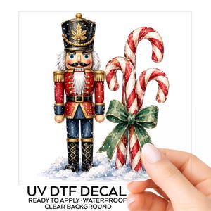 May include: A nutcracker figure wearing a black hat with a gold leaf emblem, a red and blue uniform, and white hair, standing next to two red and white striped candy canes tied with a green bow, all set against a snowy base with text below.
