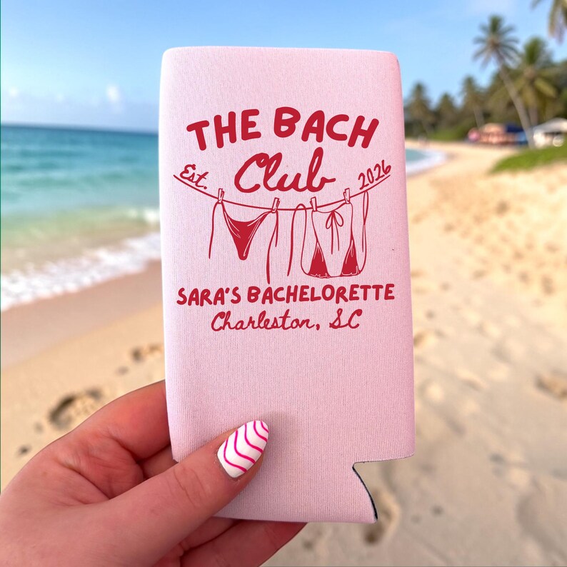 May include: A light pink foam can cooler featuring red text that reads THE BACH CLUB Est. 2026, SARA'S BACHELORETTE, Charleston, SC, with a graphic of two bikinis hanging on a clothesline between the text elements.