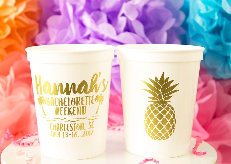 May include: Two white plastic cups with gold lettering. One cup says "Hannah's Bachelorette Weekend Charleston, SC July 13-16, 2017". The other cup has a gold pineapple design.