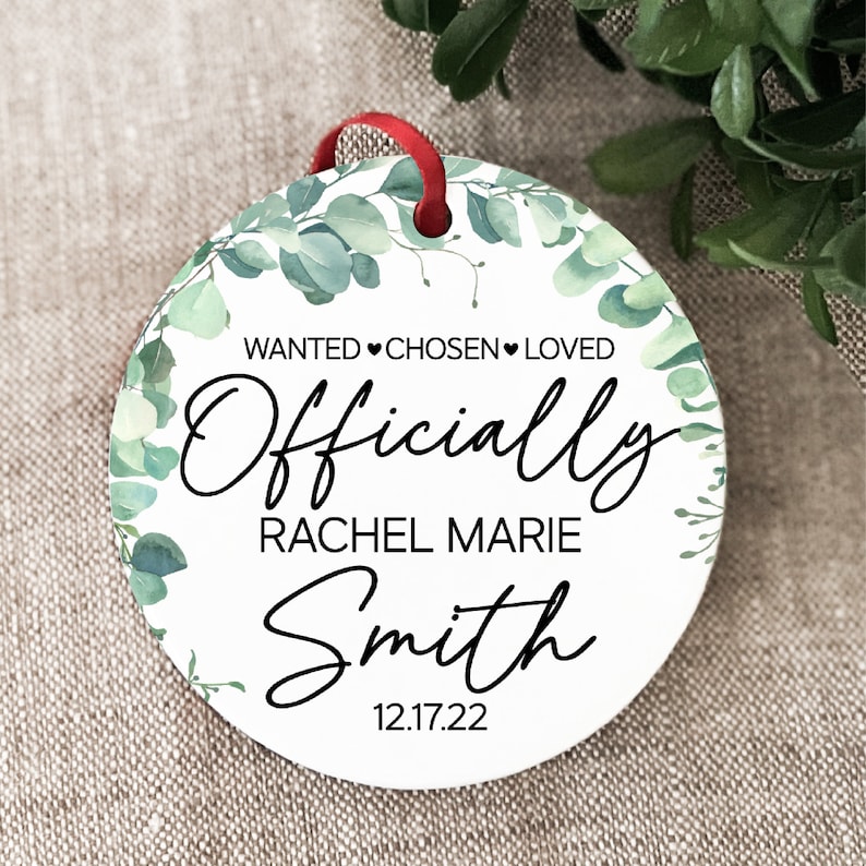Op de afbeelding: Ronde witte ornament met een groene bladrand. De ornament heeft zwarte tekst die "WANTED &hearts; CHOSEN &hearts; LOVED Officially Rachel Marie Smith 12.17.22" luidt.