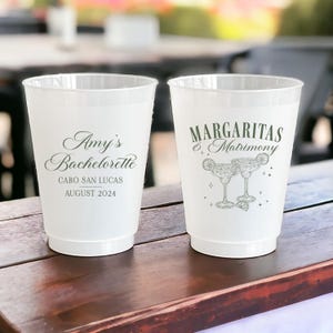 May include: Two white plastic cups with black text. The cup on the left says "Amy's Bachelorette Cabo San Lucas August 2024". The cup on the right says "Margaritas & Matrimony" with a drawing of two margarita glasses.