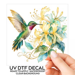 May include: A UV DTF decal featuring a watercolour illustration of a hummingbird with green, gold, and magenta feathers hovering near yellow honeysuckle flowers and green leaves. The design includes gold accents and text reading UV DTF DECAL.