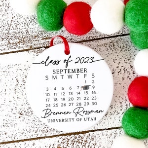 May include: White round bauble with a black calendar for September 2023. The calendar includes a graduation cap icon. The text "class of 2023" is written in cursive at the top. The text "Brennen Rossman" is written below the calendar. The text "UNIVERSITY OF UTAH" is written below the name.
