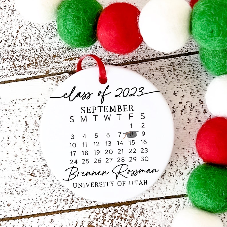 May include: White round bauble with a black calendar for September 2023. The calendar includes a graduation cap icon. The text "class of 2023" is written in cursive at the top. The text "Brennen Rossman" is written below the calendar. The text "UNIVERSITY OF UTAH" is written below the name.