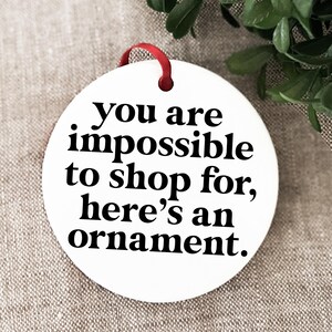 K&ouml;nnte beinhalten: Wei&szlig;er Keramik-Weihnachtsbaumschmuck mit schwarzem Text, der lautet: "Du bist unm&ouml;glich zu beschenken, hier ist ein Ornament."