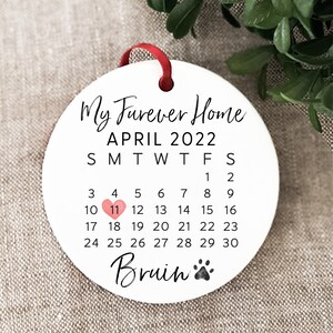 Op de afbeelding: Witte keramische ornament met de tekst "My Forever Home" en "APRIL 2022" met een kalenderrooster. De 11e is gemarkeerd met een roze hart. De tekst "Bruin" staat onderaan met een pootprint.