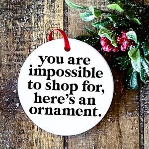 K&ouml;nnte beinhalten: Runder wei&szlig;er Weihnachtsbaumschmuck mit schwarzem Text, der lautet: "Du bist unm&ouml;glich zu beschenken, hier ist ein Ornament."