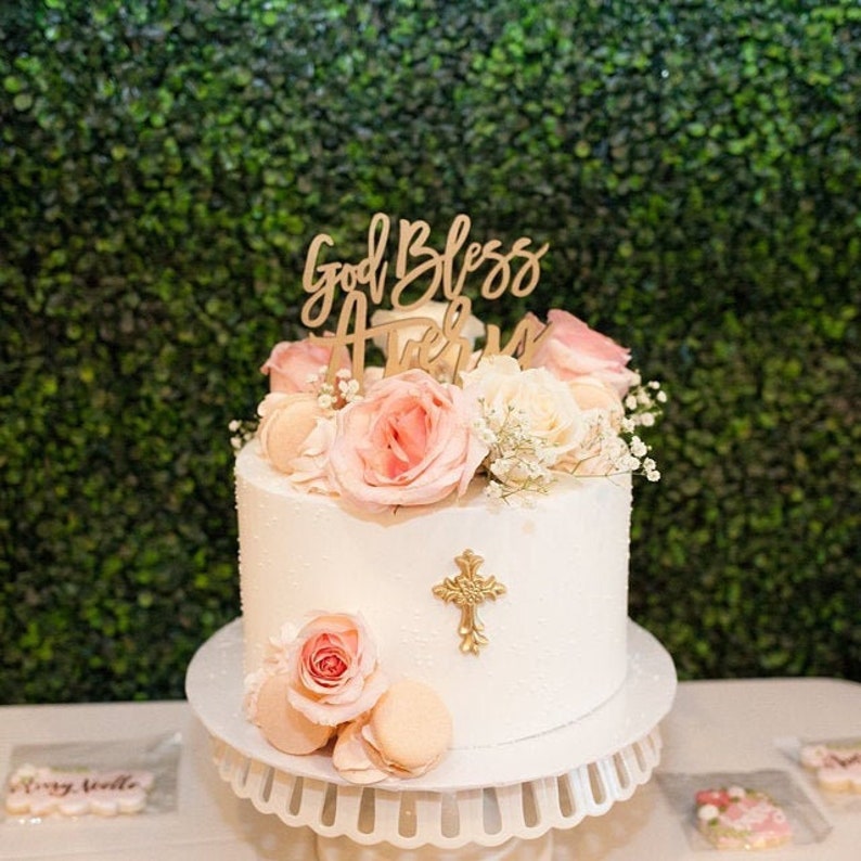 May include: A white two-tiered cake decorated with pink roses, white flowers, and macarons. The cake has a gold cross topper and a wooden sign that says "God Bless Avery".