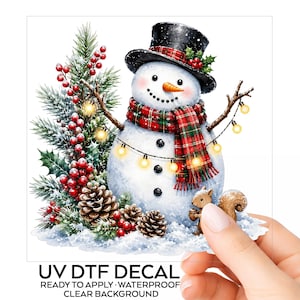 May include: A UV DTF decal featuring a snowman wearing a black top hat and a red and green plaid scarf. The snowman is decorated with a string of warm yellow lights, pine branches, red berries, pinecones, and a small brown squirrel.