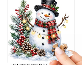 Könnte beinhalten: Ein UV-DTF-Aufkleber mit einem Schneemann, der einen schwarzen Zylinder und einen rot-grün karierten Schal trägt. Der Schneemann ist mit einer gelben Lichterkette, Tannenzweigen, roten Beeren, Tannenzapfen und einem kleinen braunen Eichhörnchen verziert.