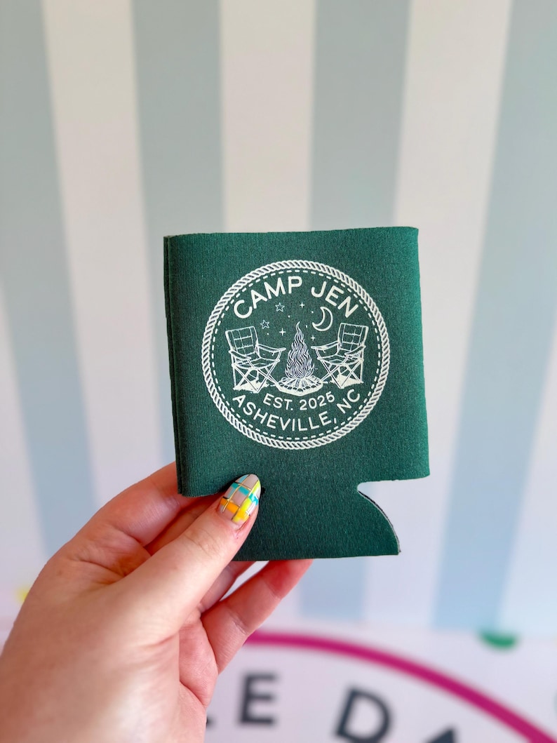 May include: A dark green foam can cooler featuring a white circular graphic with a rope border. The design includes two folding chairs, a campfire, a crescent moon, stars, and the text "CAMP JEN EST. 2025 ASHEVILLE, NC" printed in white.