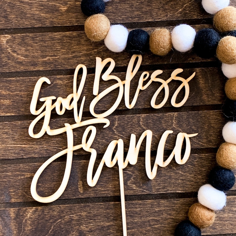 May include: Wooden cake topper with the words "God Bless Franco" written in cursive script. The topper is on a brown wooden background.