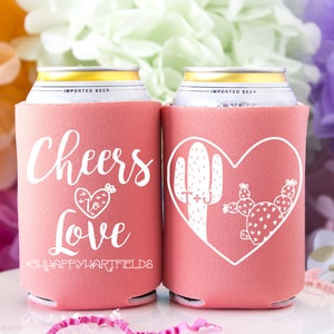 May include: Two pink can coolers with white text and designs. One cooler says "Cheers to Love" with a heart and arrow. The other cooler has a heart with two cactus plants inside. Both coolers have the hashtag "#OHAPPYHARTFIELDS".