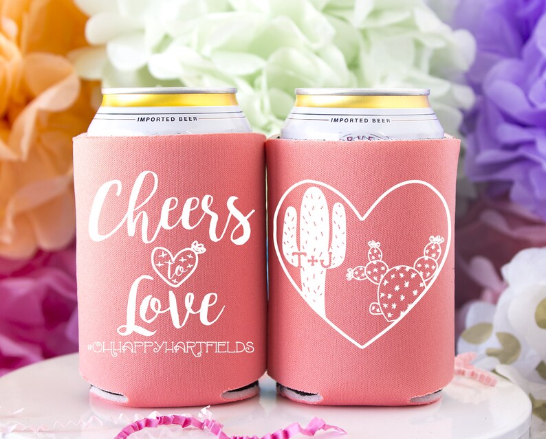 May include: Two pink can coolers with white text and designs. One cooler says "Cheers to Love" with a heart and arrow. The other cooler has a heart with two cactus plants inside. Both coolers have the hashtag "#OHAPPYHARTFIELDS".