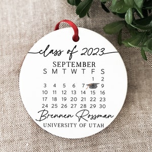 May include: White round bauble with black text that reads "class of 2023", "SEPTEMBER", "SMTWTFS", and a calendar with the dates 1-30. The bauble also includes a graduation cap icon and the text "Brennen Rossman" and "UNIVERSITY OF UTAH".