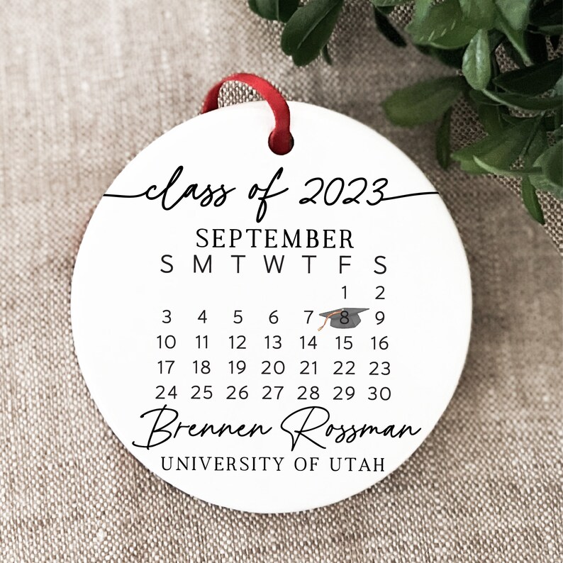 May include: White round bauble with black text that reads "class of 2023", "SEPTEMBER", "SMTWTFS", and a calendar with the dates 1-30. The bauble also includes a graduation cap icon and the text "Brennen Rossman" and "UNIVERSITY OF UTAH".