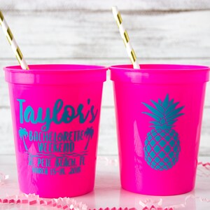 May include: Two pink plastic cups with straws. One cup has the text "Taylor's Bachelorette Weekend St. Pete Beach, FL March 15-18, 2018" printed on it. The other cup has a teal pineapple graphic.
