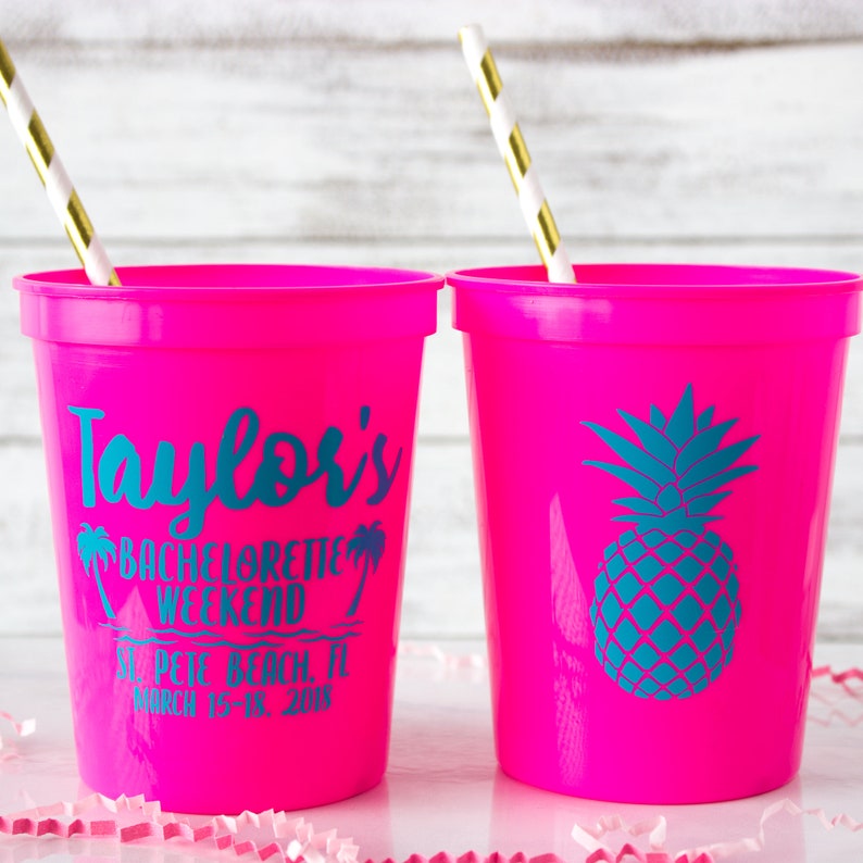 May include: Two pink plastic cups with straws. One cup has the text "Taylor's Bachelorette Weekend St. Pete Beach, FL March 15-18, 2018" printed on it. The other cup has a teal pineapple graphic.