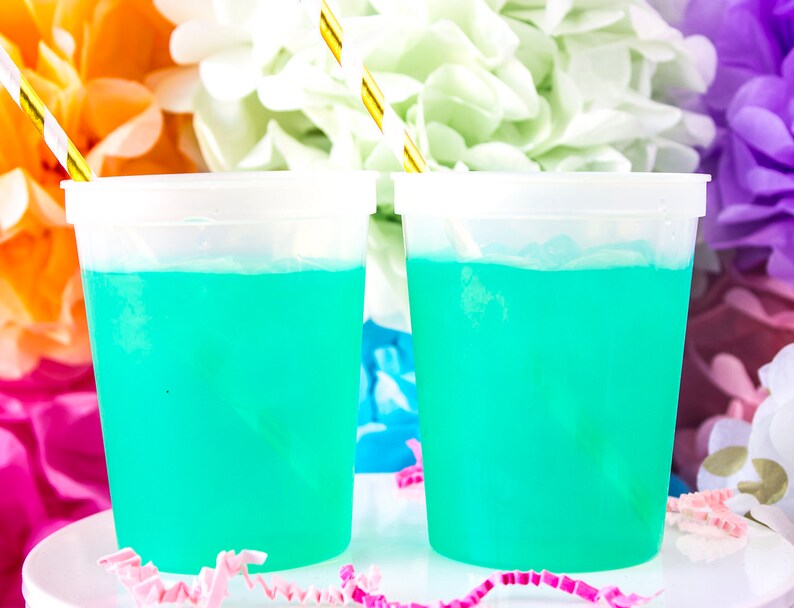 May include: Two clear plastic cups filled with a bright turquoise blue liquid. The cups are sitting on a white surface with pink and white confetti.