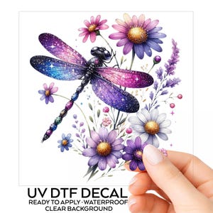 May include: A UV DTF decal featuring a galaxy-patterned dragonfly with purple, blue, and pink wings adorned with stars, surrounded by a cluster of purple, pink, and white daisy-like flowers and sprigs of lavender on a clear background.