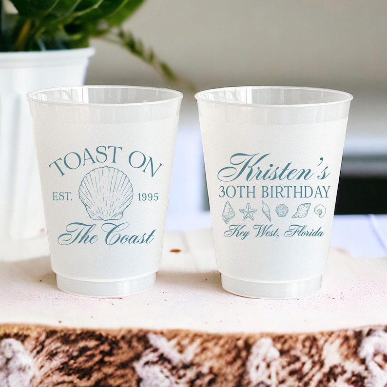 K&ouml;nnte beinhalten: Zwei wei&szlig;e Plastikbecher mit blauem Text und Bildern. Der linke Becher sagt "Toast On Est. 1995 The Coast" mit einem Muschelbild. Der rechte Becher sagt "Kristen's 30th Birthday Key West, Florida" mit einem Seestern und Muscheln.