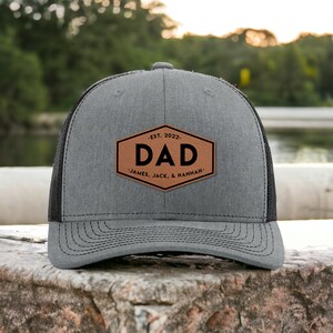 May include: A gray and black trucker hat with a brown leather patch that says "EST. 2022 DAD JAMES, JACK, & HANNAH".