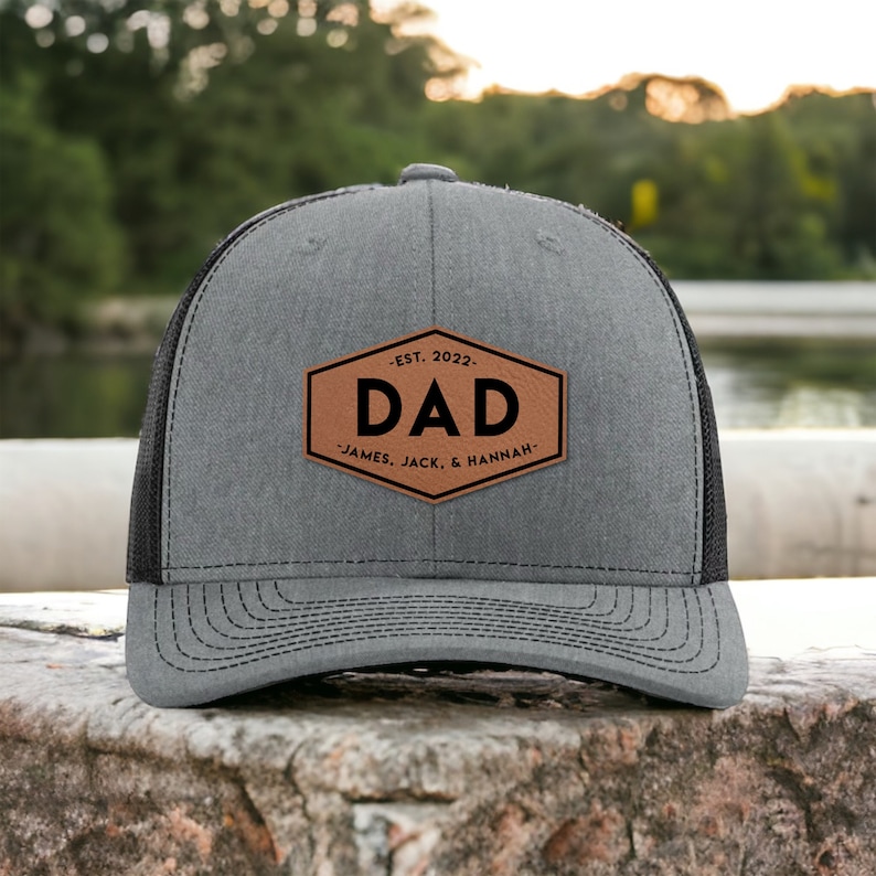 May include: A gray and black trucker hat with a brown leather patch that says "EST. 2022 DAD JAMES, JACK, & HANNAH".